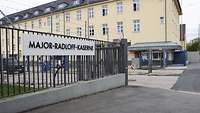 An einem Metallzaun hängt das große Kasernenschild, daneben die Einfahrt und ein Wachbereich