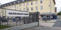 An einem Metallzaun hängt das große Kasernenschild, daneben die Einfahrt und ein Wachbereich