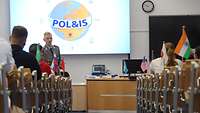 Ein Soldat steht vor sitzenden Personen. Hinter ihm ist das Logo „POL&IS“ auf eine Wand projeziert.
