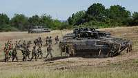 Mehrere bewaffnete Soldaten laufen hinter zwei fahrenden Panzern vom Typ Puma im Gelände.