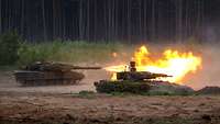 Ein Panzer vom Typ Leopard 2 schießt scharf im Gelände, schräg davor fährt ein Panzer vom Typ Puma.