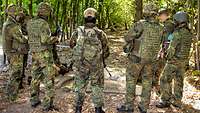 Mehrere Soldaten stehen zur Lagebesprechung im Kreis zusammen und schauen auf den Waldboden.