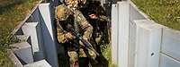 Ein Soldat mit Waffe im Anschlag geht durch einen Graben, auf dem ein Gitter mit Zweigen und Blättern liegt.