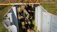 Ein Soldat mit Waffe im Anschlag geht durch einen Graben, auf dem ein Gitter mit Zweigen und Blättern liegt.