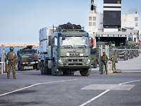 aus einem Tor, bewacht von Soldaten, kommen große Militärfahrzeuge aus einem Frachter