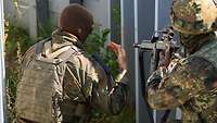 A soldier with his weapon at the ready stands behind a soldier who is explaining something to him.