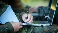 Die Hand eines Soldaten am Laptop, gleichzeitig schreibt er mit Block und Stift mit.