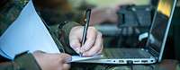 Die Hand eines Soldaten am Laptop, gleichzeitig schreibt er mit Block und Stift mit.