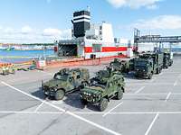 Etwa zehn deutsche und schwedische Militärfahrzeuge stehen in einem Hafen, im Hintergrund ein Schiff mit geöffneter Laderampe