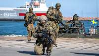 Drei Soldaten im Hafen, alle haben Waffen in der Hand. Im Hintergrund ein dunkelgrünes Boot und ein Schiff