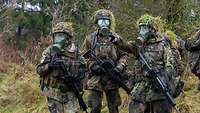 Drei Soldaten stehen mit aufgesetzter ABC-Schutzmaske in einem Waldstück.