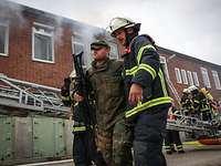 Zwei Feuerwehrleute tragen einen verwundeten Soldaten aus einem brennenden Haus.