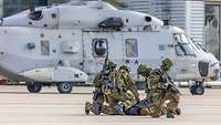 Soldaten drücken eine Person zu Boden bei einer Übung. Im Hintergrund steht ein Hubschrauber.