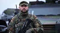 Ein Soldat steht vor einem grünen Militärfahrzeug. In der Hand trägt er ein Gewehr. 