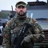 Ein Soldat steht vor einem grünen Militärfahrzeug. In der Hand trägt er ein Gewehr. 