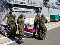 Personen in militärischer Schutzausrüstung tragen eine rote Trage von Bord eines Schiffes