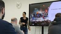 Lehrkraft steht vor einem Smartboard und spricht zu Schülerinnen und Schülern.