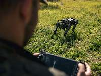 Ein Soldat steht mit einer Fernbedienung auf einer Wiese hinter einem vierbeinigen Roboterhund.