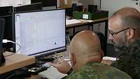 Zwei Soldaten sitzen in Uniform in einem Büro vor einem Monitor und passen den Quellcode einer Software an.