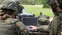 Eine Soldatin und ein Soldaten stehen im Gelände an einem Laptop und laden Software auf ein Gerät.