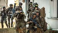 Bewaffnete Soldaten laufen in einer Reihe an einer Hauswand entlang, einige hocken im Vordergrund.