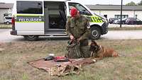 Ein Soldat kniet auf einer ausgebreiteten Decke auf einer Wiese. Neben ihm sitzt ein Hund.