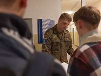 Ein Soldat im Feldanzug blickt auf Unterlagen. Zwei Männer in ziviler Kleidung stehen ihm gegenüber.