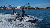 Zwei Soldaten fahren auf Jetski ähnlichen Wasserfahrzeugen. Hinten ist ein Hafen mit Werft zu sehen.