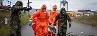 Fünf Personen in orangen und militärischen Schutzanzügen tragen einen Menschen.