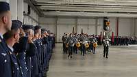 Militärmusiker marschieren durch eine Flugzeughalle. Links stehen Luftwaffensoldaten in einer Reihe.