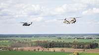 Zwei Hubschrauber fliegen hintereinander über Felder und Baumreihen.