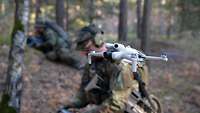 Eine Drohne schwebt durch den Wald. Im Hintergrund ist der Drohnenbediener zu erkennen.