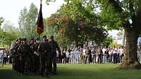 Zivilisten stehen am Rande einer Wiese. Vor ihnen marschiert eine Formation mit Truppenfahne ein.