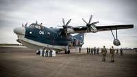 Ein blau-weißes Propeller-Flugzeug auf einem Flugplatz. Soldaten stehen drumherum.