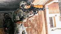 Ein Soldat steht in einem alten Haus an der geöffneten Tür. Seine Waffe zeigt nach draußen.