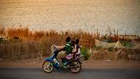 Ein Mann und zwei Mädchen fahren auf einem blauen Moped am Nigerufer auf einer Straße