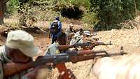 Malische Soldaten mit Waffen im Anschlag nebeneinander suchen Deckung hinter einem Hügel, im Hintergrund ein deutscher Soldat
