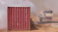 Brennende Container mit zwei Feuerwehrmännern im Rauch und ein Löschfahrzeug
