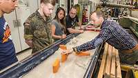 Studenten beobachten einen Versuchsaufbau im Institut für Bauingenieurwesen und Umweltwissenschaften an der Universität der Bundeswehr in München. Studenten stehen in einer Werkhalle an einem Versuchsmodell