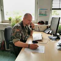 Ein Mann sitzt am Schreibtisch vor einem Monitor, Telefonhörer in der linken Hand, Schreibzeug rechts.
