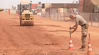 Ein Soldat auf der Straße mit einem Hammer und befestigt Pylonen im Sand. Im Hintergrund ist eine Planierraupe zusehen