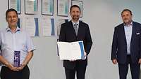 Drei männliche Person präsentieren eine Urkunde und eine Trophäe unter Wahrung der Abstandsregeln