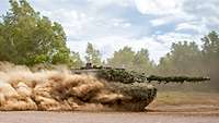 Half-enveloped in a cloud of dust, a tank speeds across a sandy training ground.