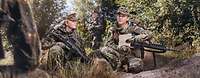Two servicewomen crouch in wooded terrain with their weapons, conferring with each other.