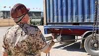 Im Vordergrund steht ein Soldat und blickt nach oben, im Hintergrund wird ein Container auf Sattelschlepper verladen