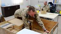Ein Soldat ordnet die Pakete in einer großen Holzkiste. Ein weiterer Soldat sitzt am Schreibtisch und hält ein Paket in der Hand