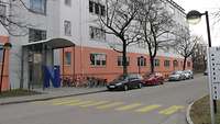 Ein großes fünfstöckiges Schulgebäude von außen. Davor parken Autos.