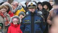Ein Marineoffizierin mit schwerer blauer Bordjacke und weißer Tellermütze steht vor einer Menschenmenge.