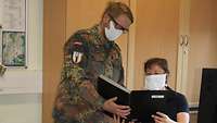 Winkler reicht Meunier ein Auftragsbuch. Meunier sitzt an ihrem Schreibtisch, Winkler steht daneben. Beide trage Mundschutz.