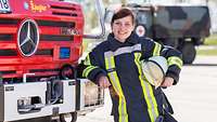 Eine Feuerwehrfrau in Uniform und mit Helm in der Hand lehnt an einem Feuerwehrfahrzeug